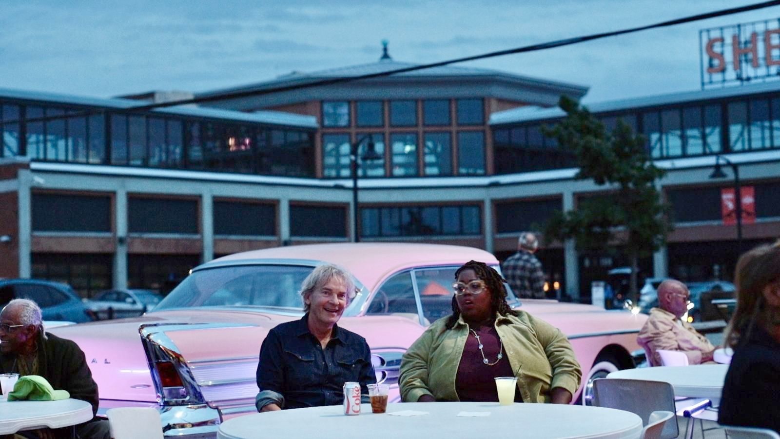 Twee mensen aan een tafel buiten bij schemering, met een roze klassieke auto erachter