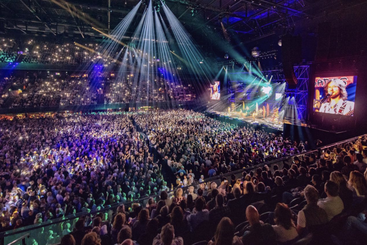 Overzicht van een volle arena met publiek en een lichtshow rond het podium